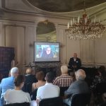 predstavenie knihy, popredseda pred publikom, na obrazovke za ním je autor knihy pripojený naživo